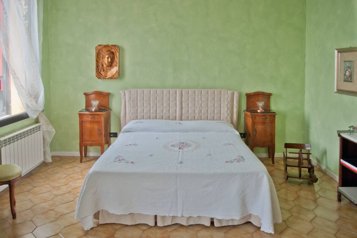 A bedroom with a bed, two nightstands, a chair, a radiator, and a window with curtains.