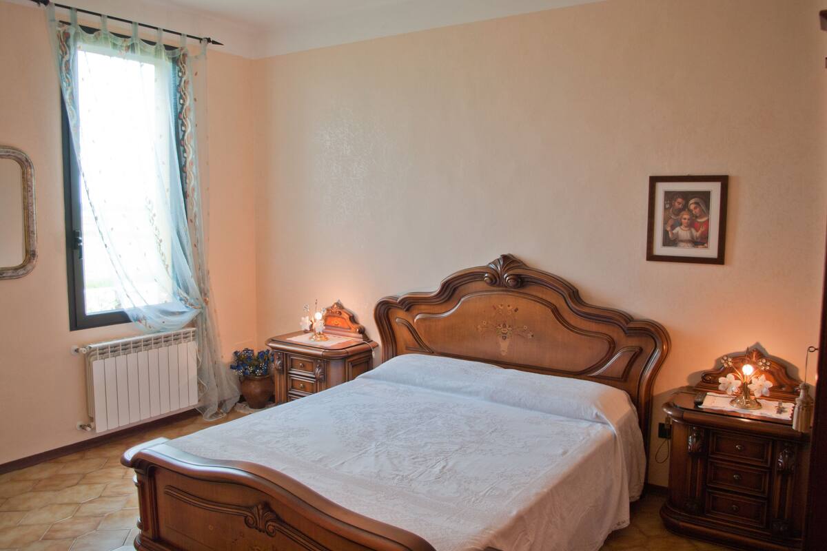 A bedroom with a wooden bed, a bedside table with a lamp, a radiator, and a window with curtains.