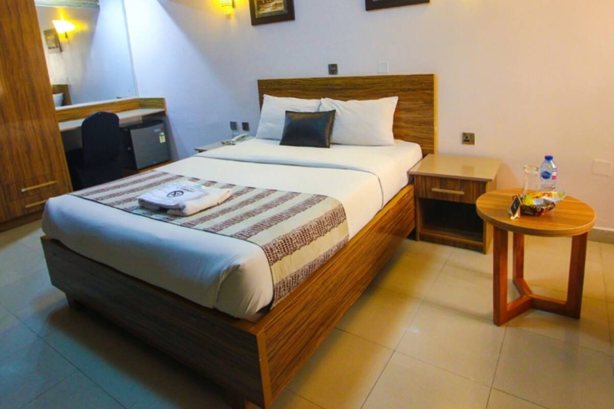 A hotel room with a wooden bed, a desk, and a small table with a water bottle.