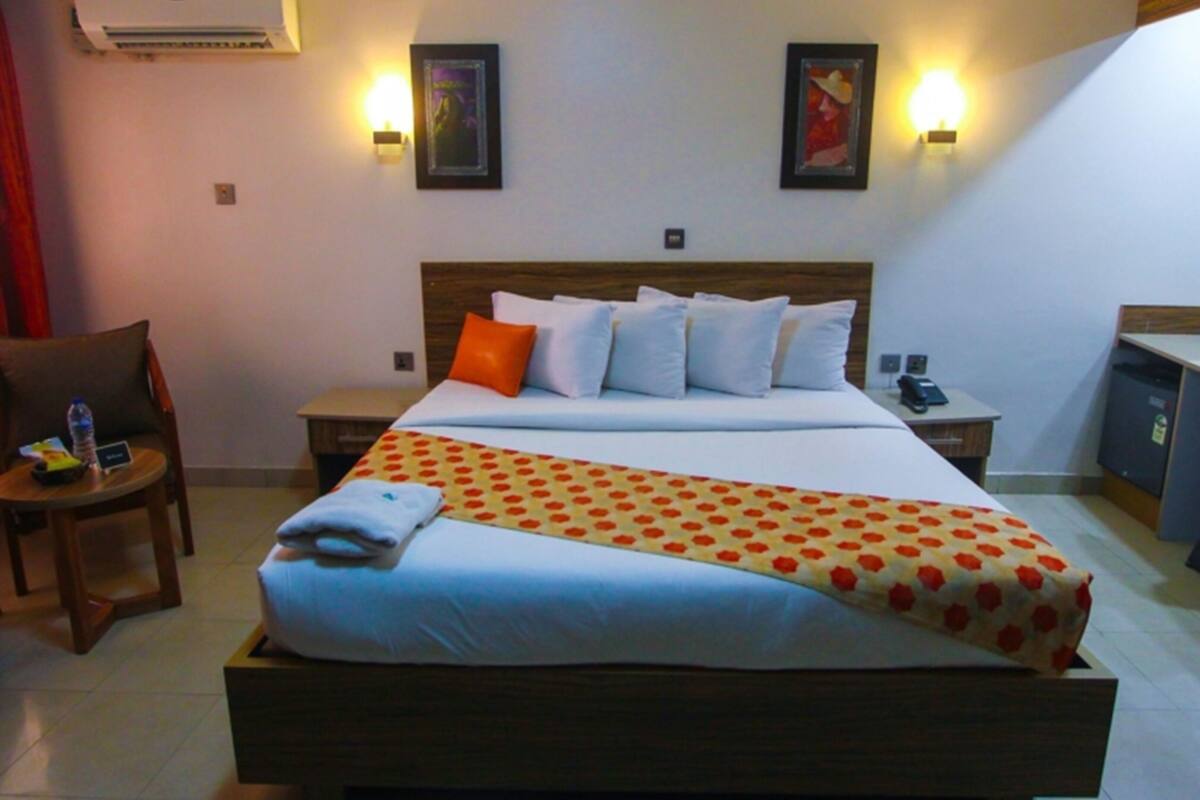 A hotel room with a large bed, two bedside tables, a chair, an air conditioning unit, and two wall-mounted lights.