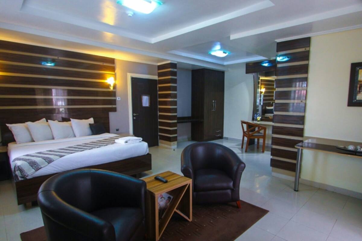 A modern hotel room with a large bed, two leather armchairs, a small wooden table, and a desk with a chair.