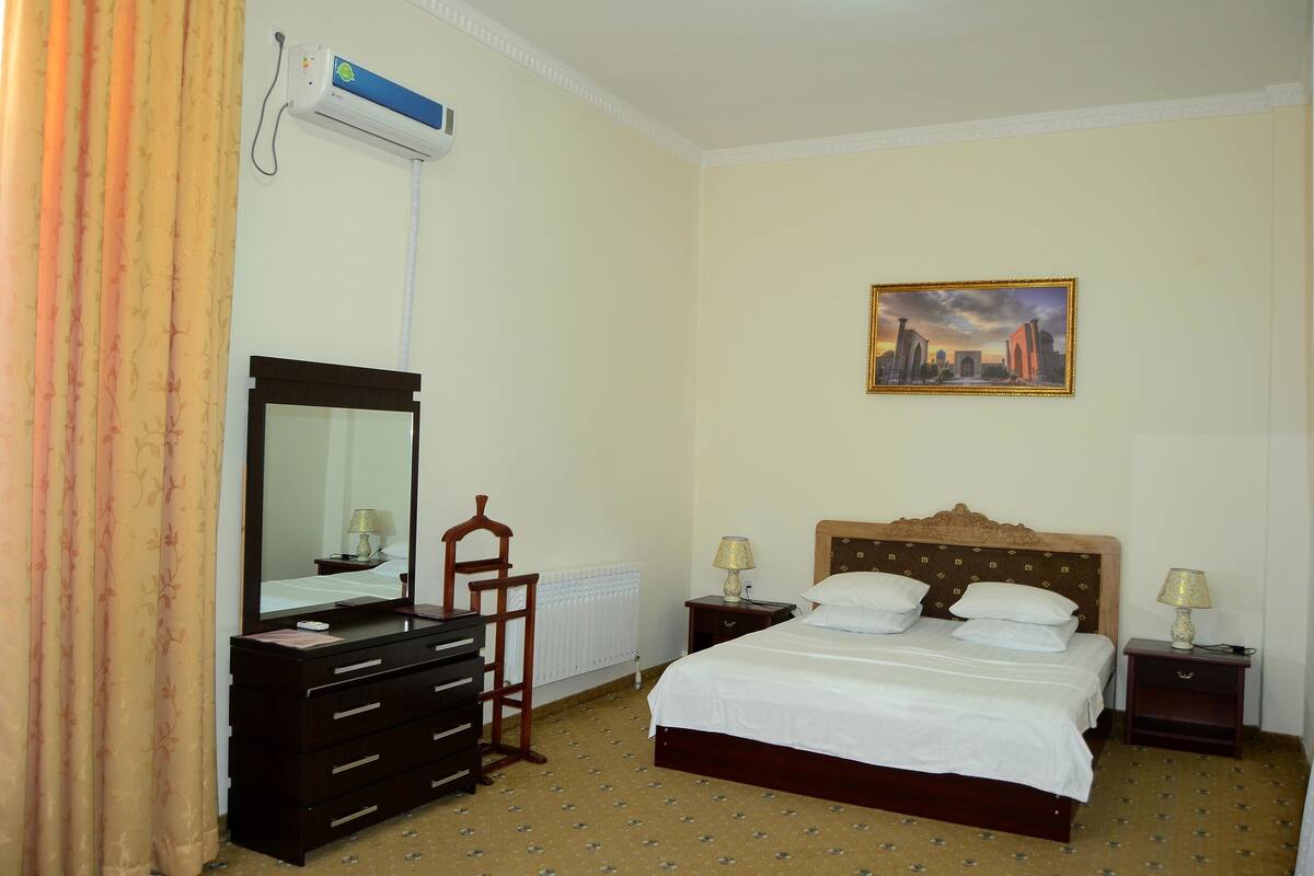 A hotel room with a bed, a dresser, a mirror, a painting on the wall, and an air conditioning unit.