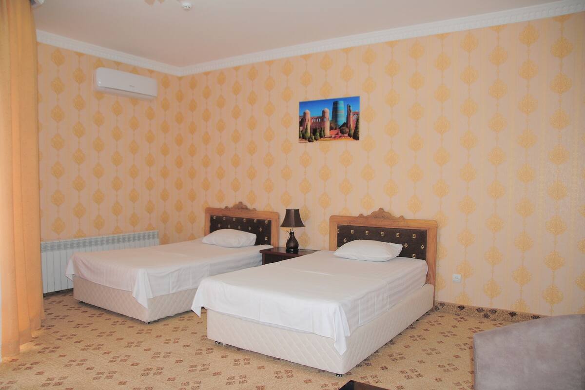 A hotel room with two single beds, a nightstand with a lamp, a wall-mounted air conditioner, and a patterned wallpaper.