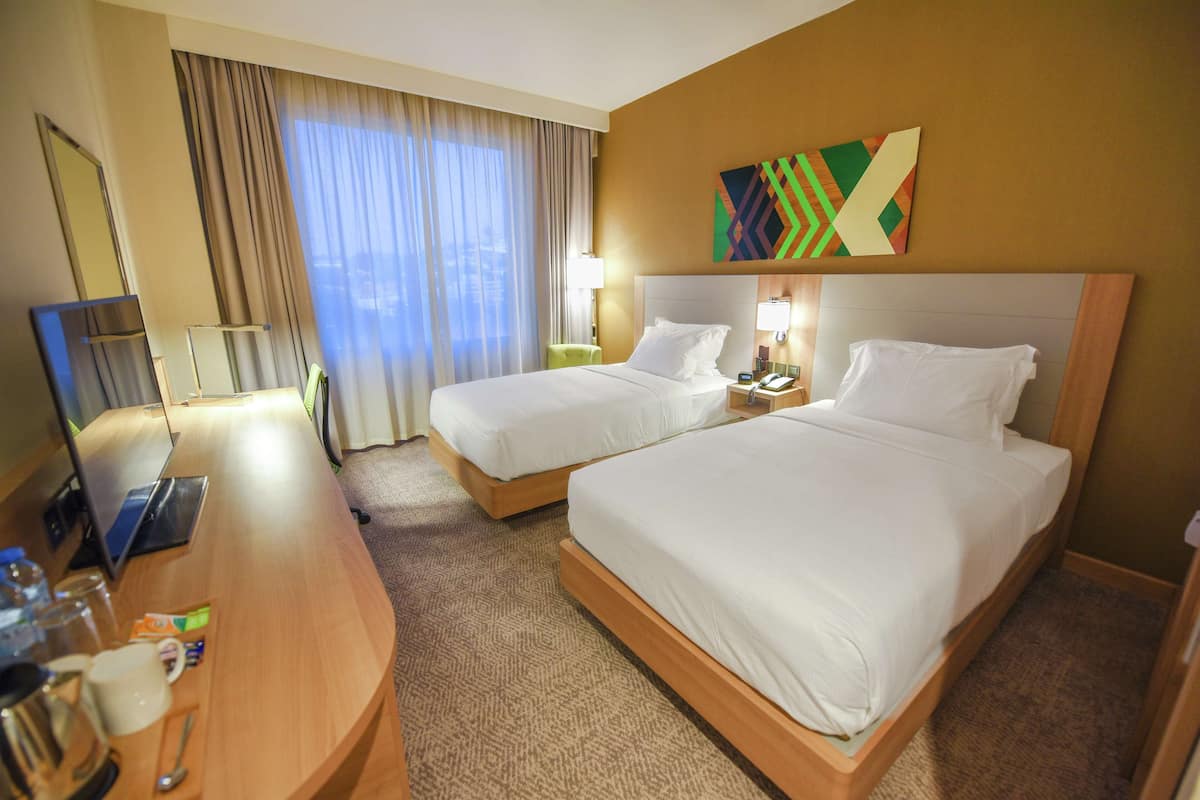 Une chambre d’hôtel avec deux lits, un bureau, un miroir et une fenêtre avec des rideaux.