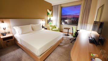 A hotel room with a large bed, a desk, a chair, a TV, and a window with a city view.