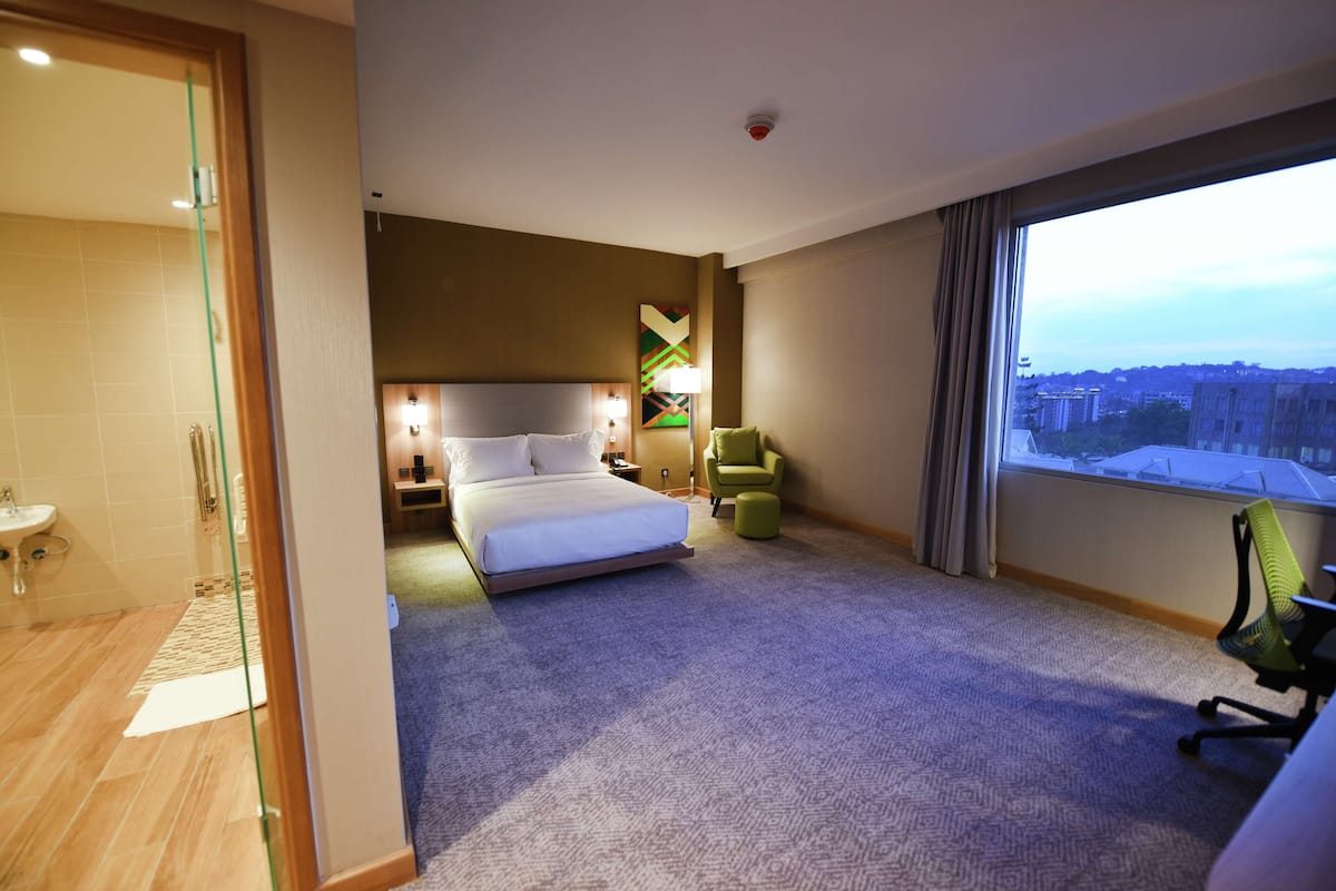 Une chambre d’hôtel comprenant un lit, un bureau, une chaise, une fenêtre donnant sur la ville et une salle de bain avec une porte en verre.
