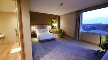 A hotel room with a bed, a desk, a chair, a window with a city view, and a bathroom with a glass door.