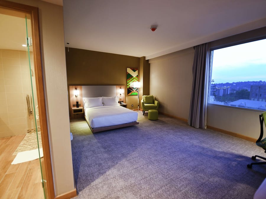 Habitación de hotel con cama, escritorio, silla, ventana con vistas a la ciudad y baño con puerta de vidrio.