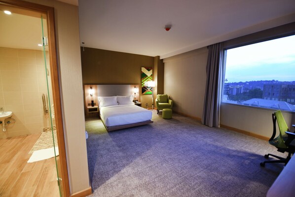 Habitación de hotel con cama, escritorio, silla, ventana con vistas a la ciudad y baño con puerta de vidrio.
