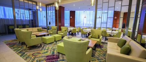 A modern hotel lobby with green seating, a patterned carpet, and large windows.