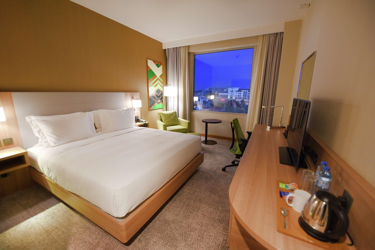 Habitación de hotel con cama, mesita de noche, lámpara, silla, pequeña mesa y vista a la ciudad de noche.
