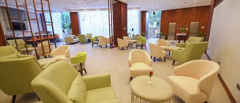 Un moderno vestíbulo de hotel con sillones verde y beige, una mesa redonda central con un florero, y una zona de cocina visible al fondo.