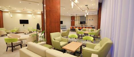 Un vestíbulo de hotel con varias zonas de estar, incluyendo sofás y sillones, un mostrador de recepción y una pantalla de televisión.