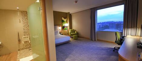 A hotel room with a bed, a desk, a chair, a large window with a city view, and a shower area.