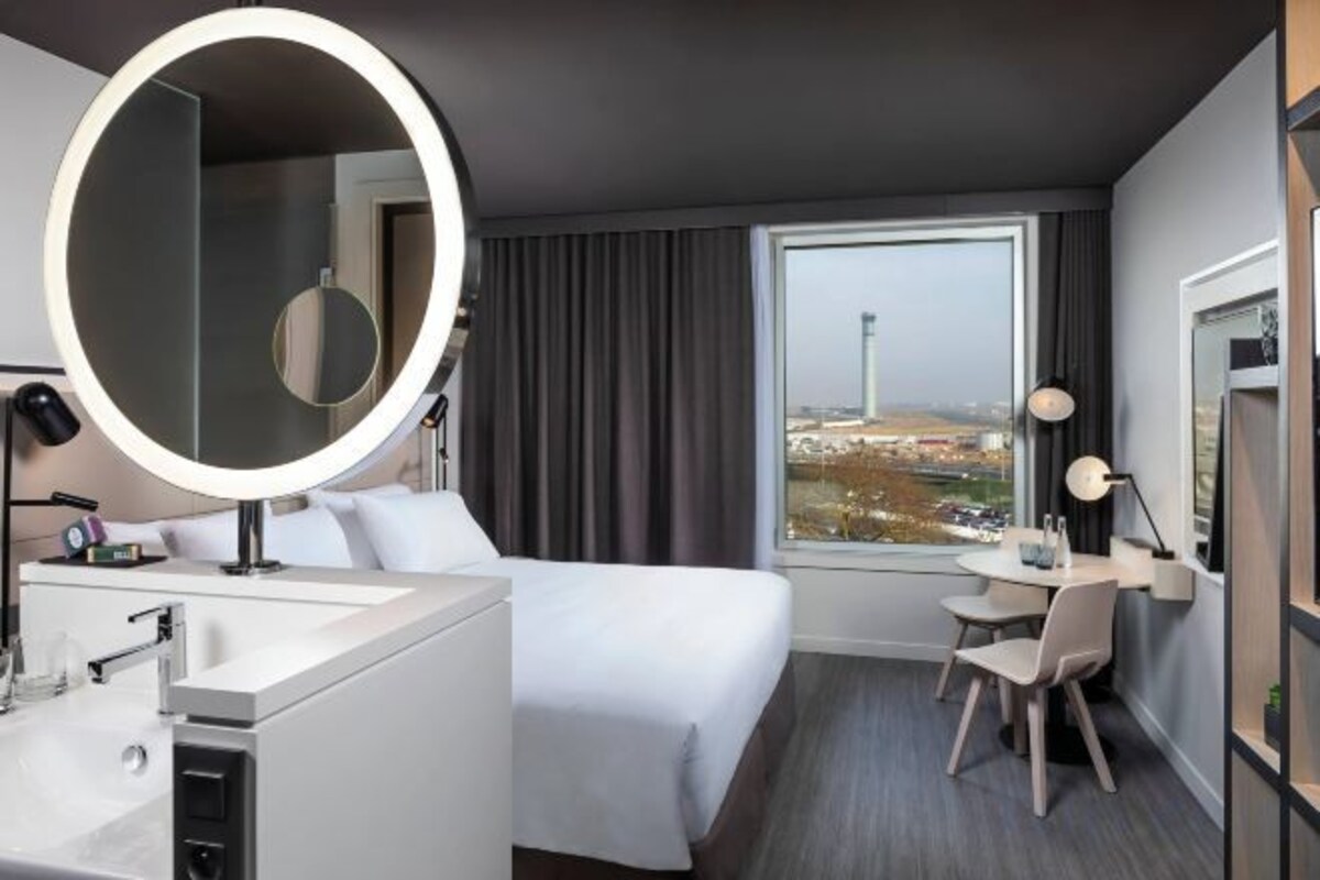 Habitación de hotel con cama, escritorio, silla, espejo redondo y vistas a un paisaje urbano a través de un ventanal grande.