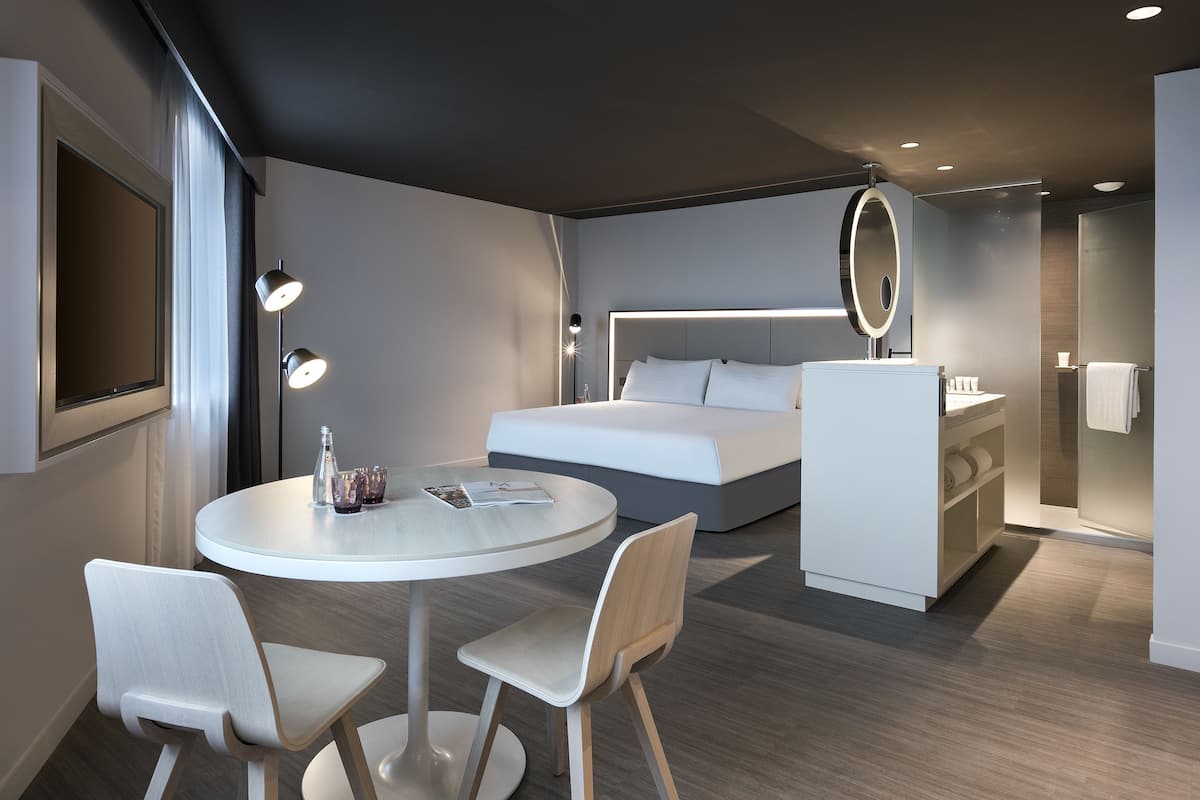 A modern hotel room with a round dining table, two chairs, a bed with a headboard mirror, a built-in wardrobe, and a small bathroom.
