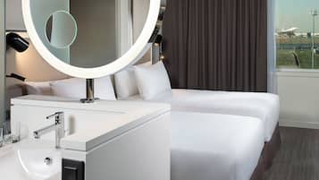 Una habitación de hotel moderna con un espejo redondo, una cama blanca, una mesita de noche con lámpara y vistas al aeropuerto.