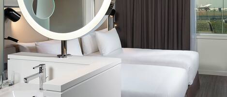Ein modernes Hotelzimmer mit einem runden Spiegel, einem weißen Bett, einem Nachttisch mit Lampe und Blick auf einen Flughafen.