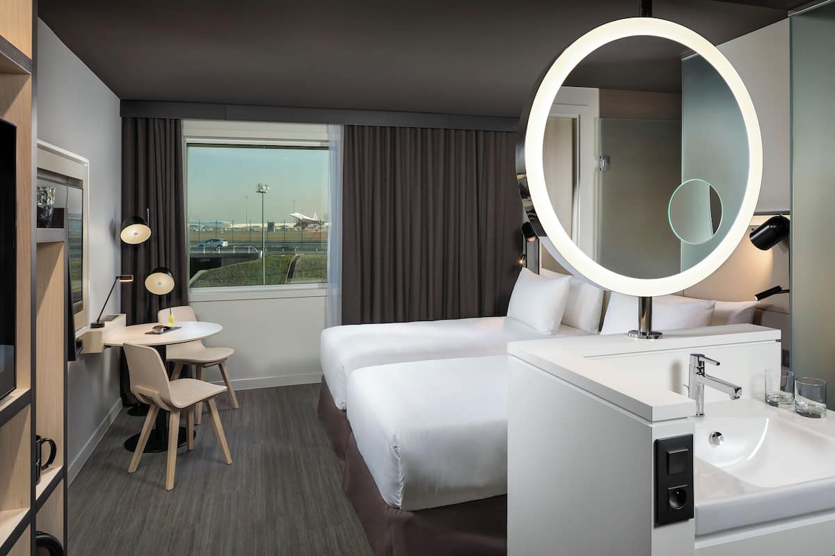 A hotel room with a bed, a desk with two chairs, a round mirror, a white vanity with a sink, and a window with curtains.
