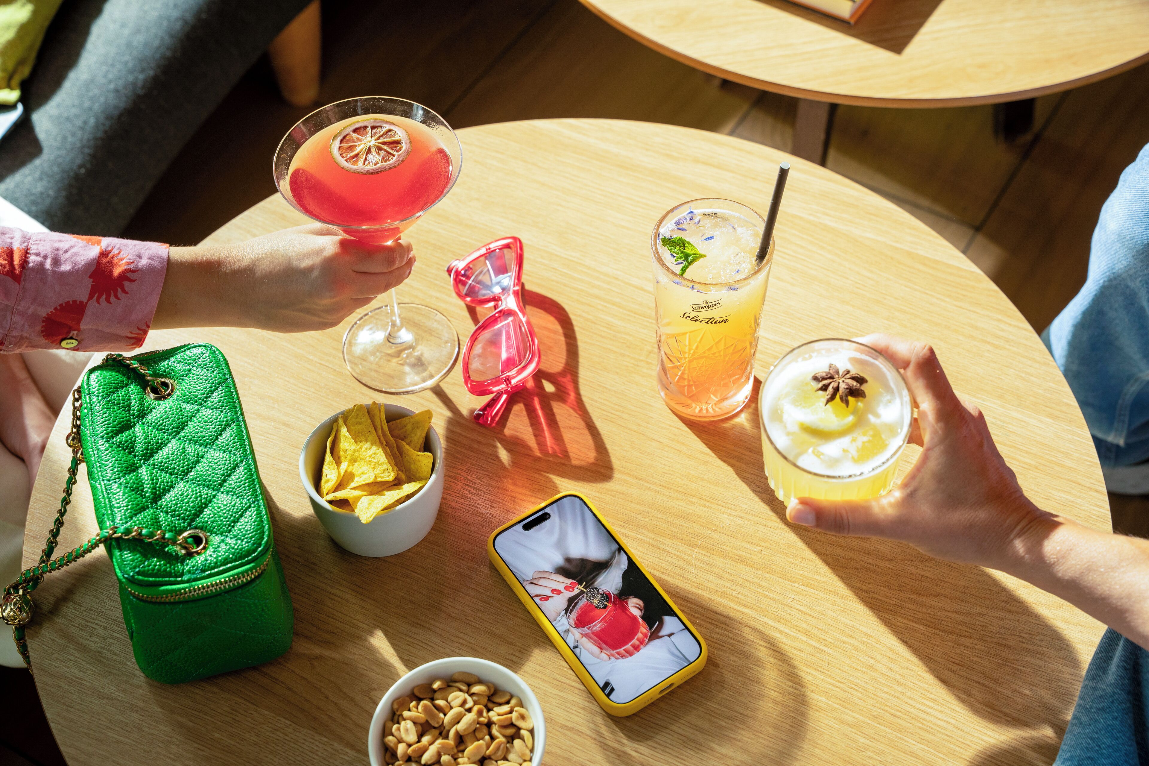 A round wooden table with various drinks, a green handbag, a smartphone, and some snacks.