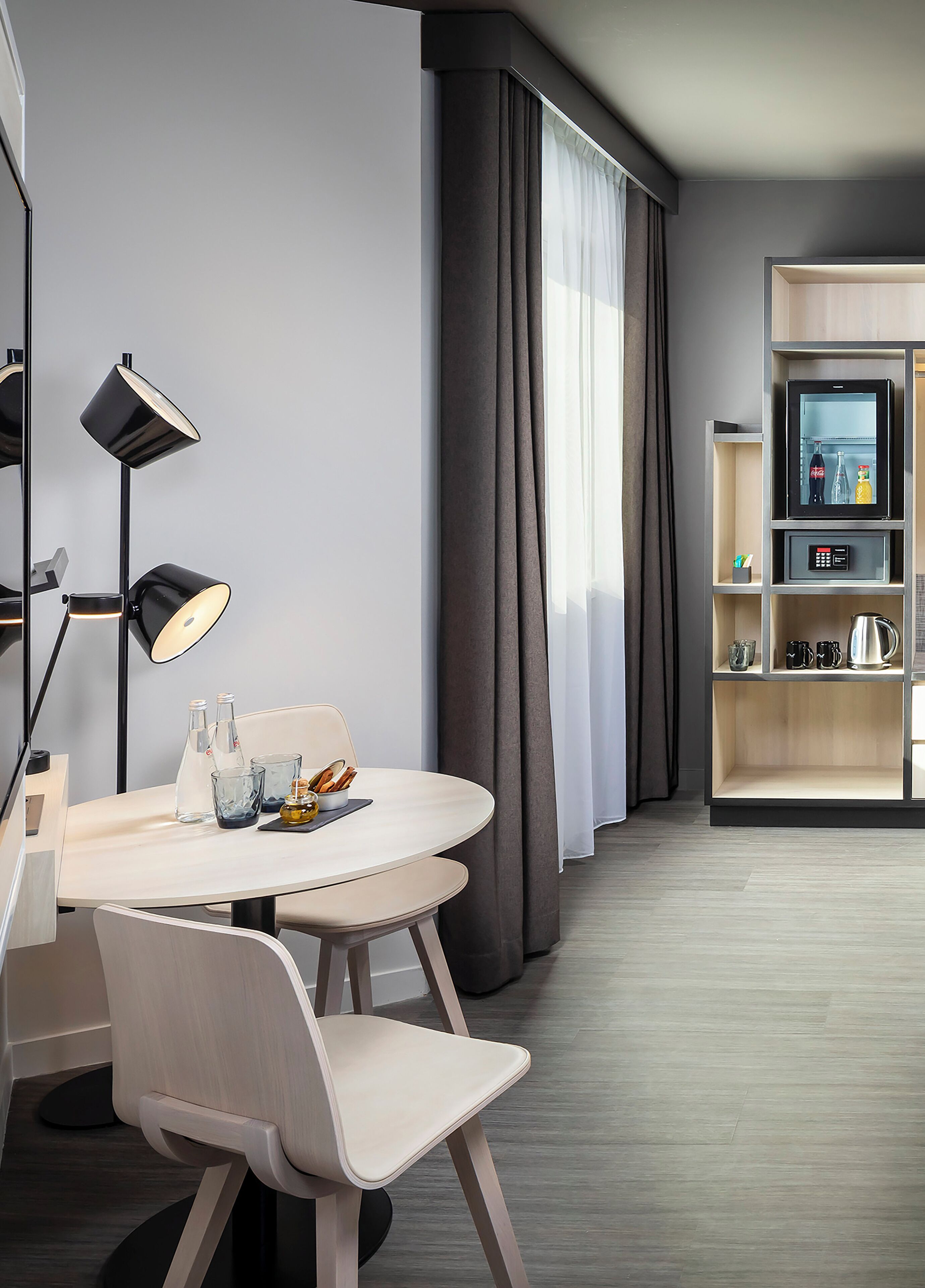 A modern hotel room with a round dining table, two chairs, a floor lamp, a minibar, and a large window with curtains.