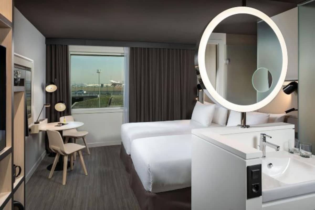 A hotel room with a bed, a desk with chairs, a round mirror, a sink, and a window with curtains.