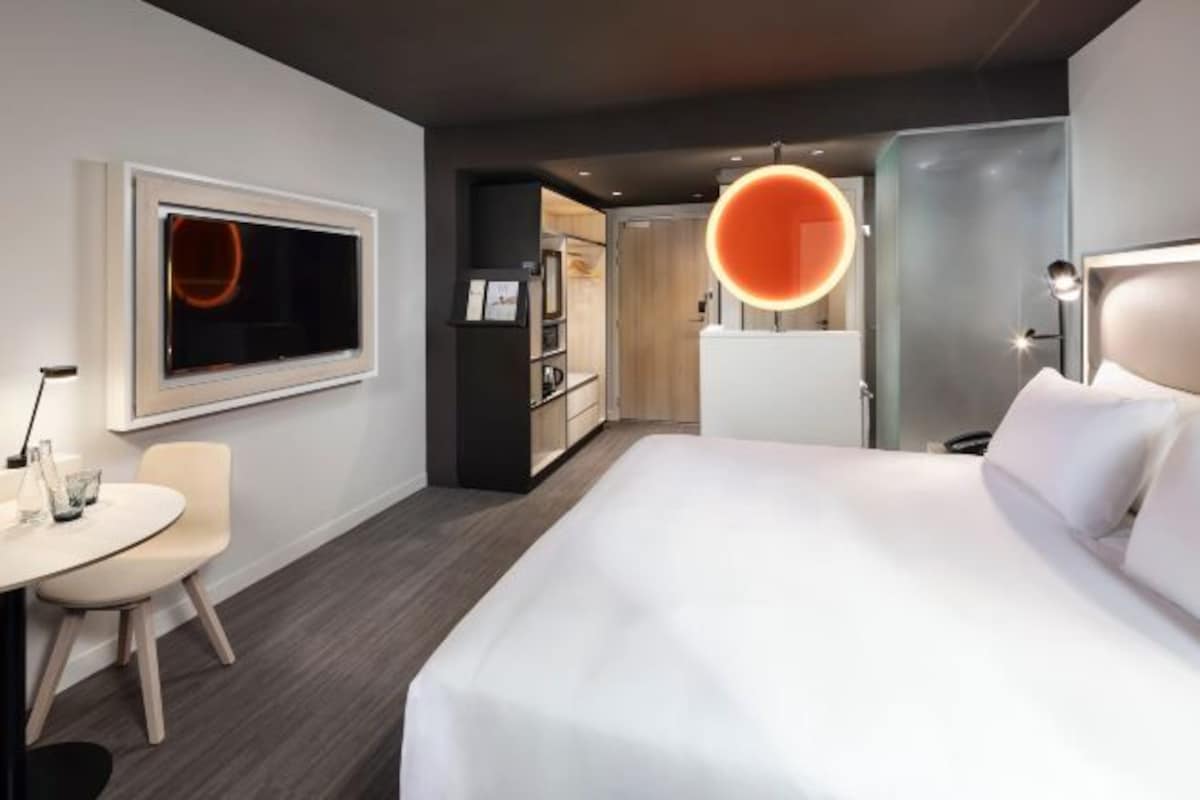 A modern hotel room with a large bed, a desk, a chair, a wardrobe, and a circular light fixture.
