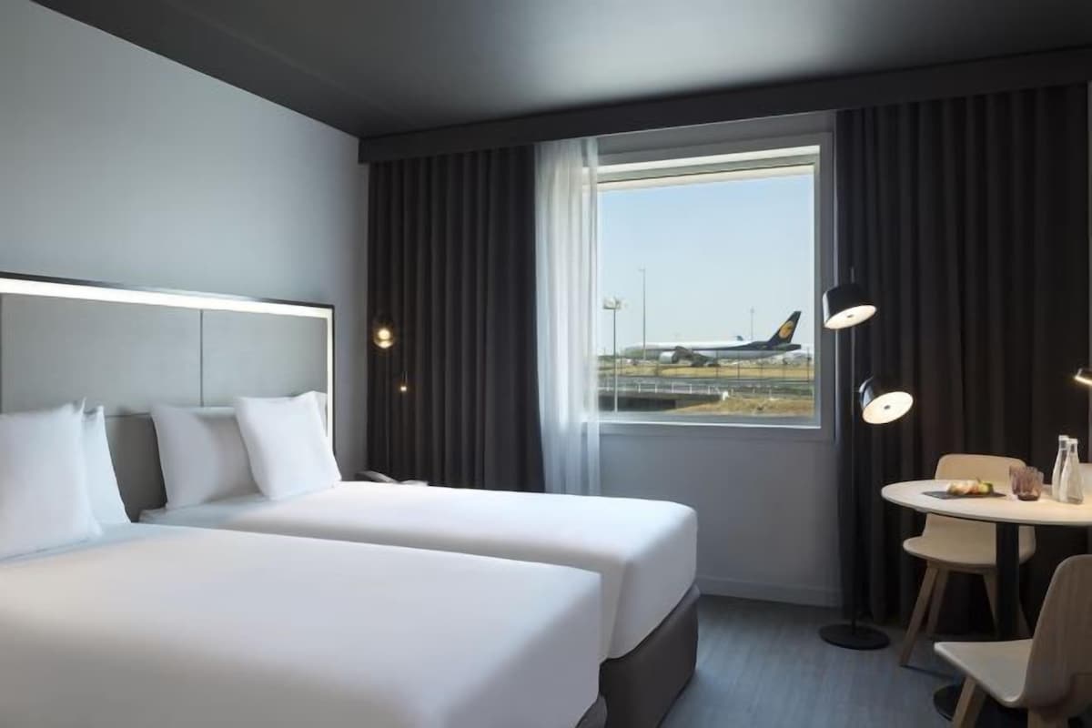 Hotellihuone, jossa on kaksi sänkyä, ikkuna, josta on näkymä lentokoneeseen, sekä pieni pöytä ja tuolit.