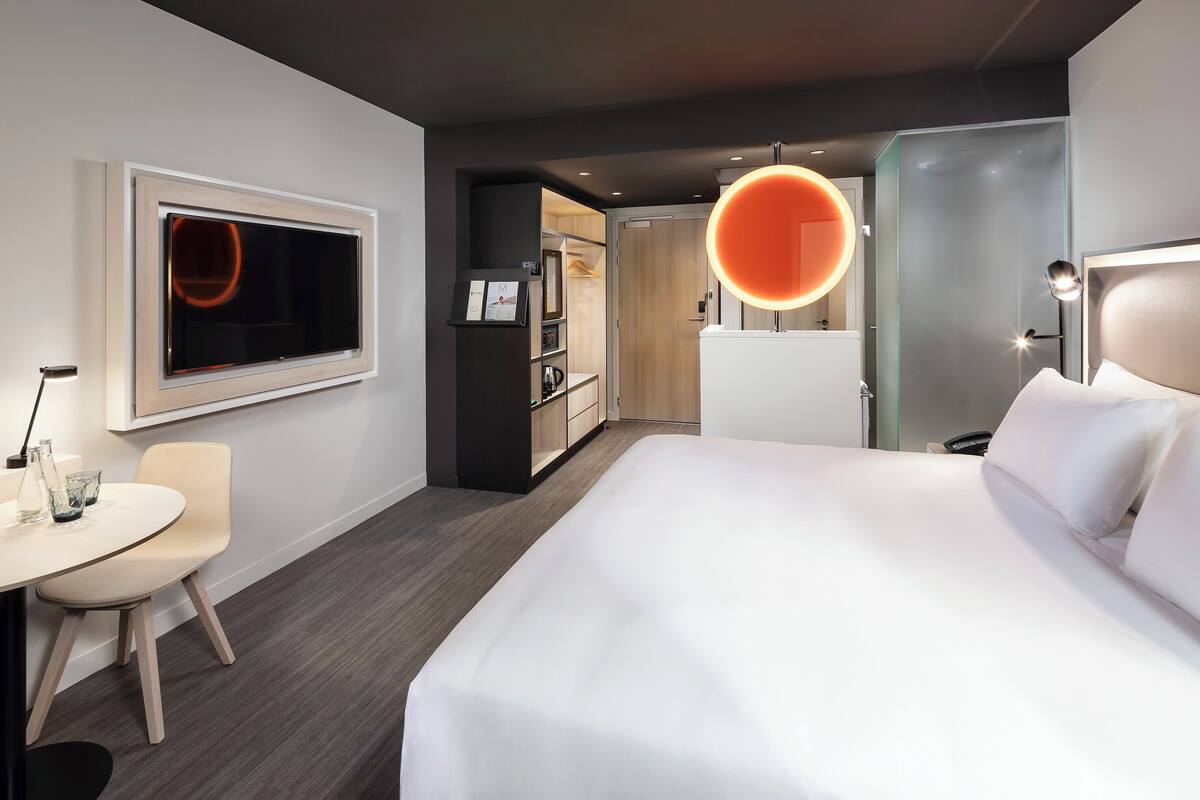 A modern hotel room with a large bed, a desk with a chair, a TV, and a circular light fixture.