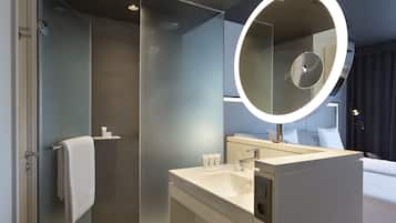 A modern bathroom with a glass shower enclosure, a large mirror with a circular light, and a white sink with toiletries.