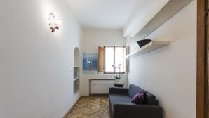 Un espacio habitable compacto con suelo de espiga, un sofá oscuro, una pequeña mesa con una planta y una ventana que ofrece vistas al exterior.