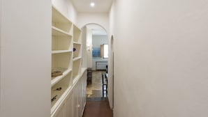 Un pasillo estrecho con paredes blancas y suelo de espiga. Estanterías a lo largo de un lado, y una cocina visible en la distancia.