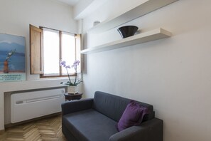 Un salon moderne avec un canapé gris, un coussin violet, une plante en pot, une fenêtre donnant sur les montagnes et une étagère murale sur laquelle est posé un objet noir.