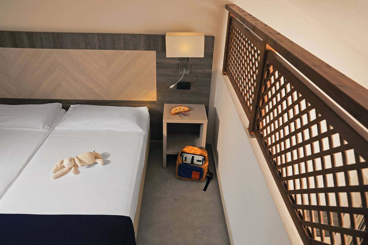 A hotel room with a bed, a nightstand with a lamp, a small table with a snack, and a wooden railing.
