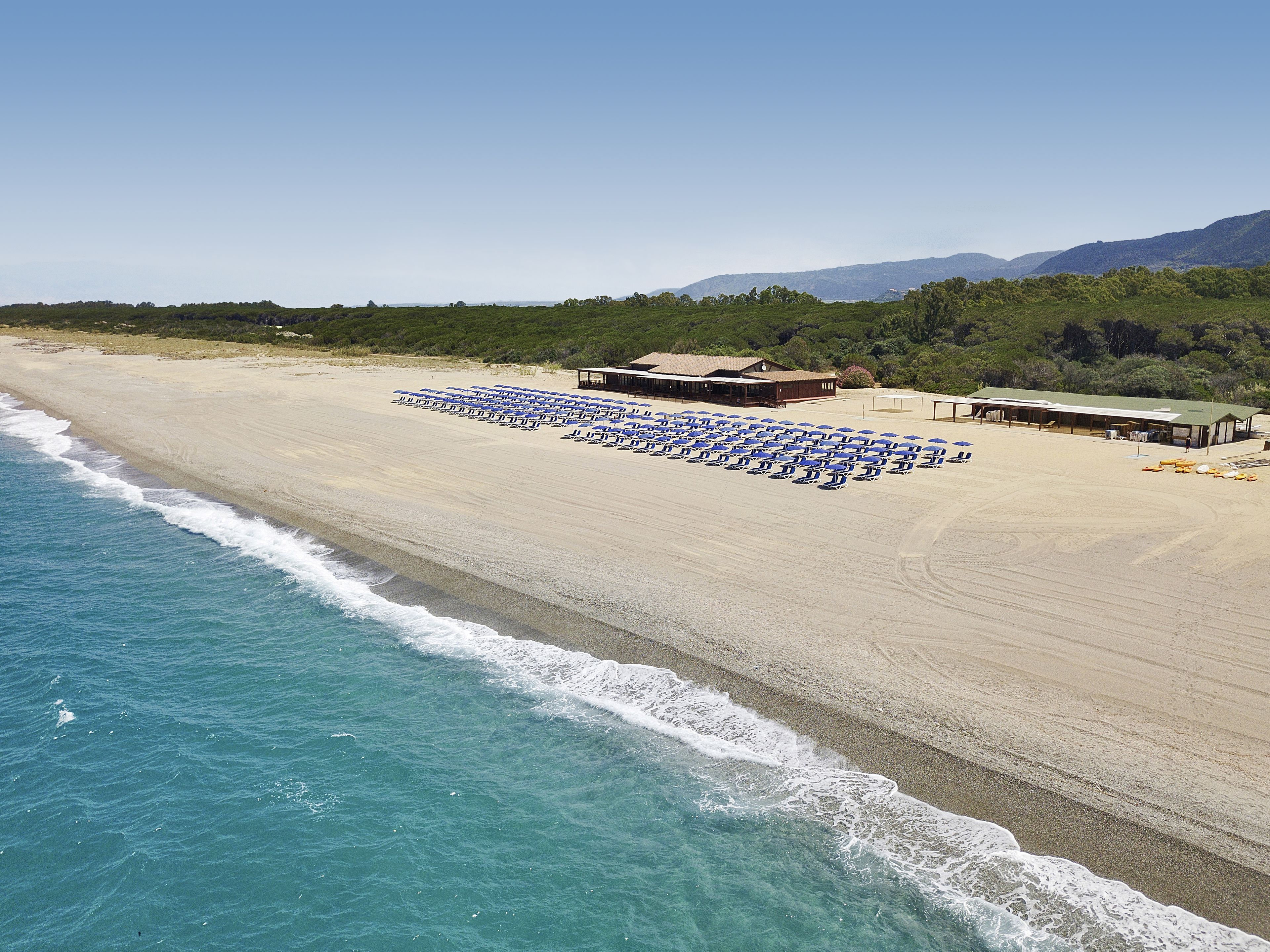 Una spiaggia con file di ombrelloni blu e sdraie, un edificio con tetto verde e montagne sullo sfondo.