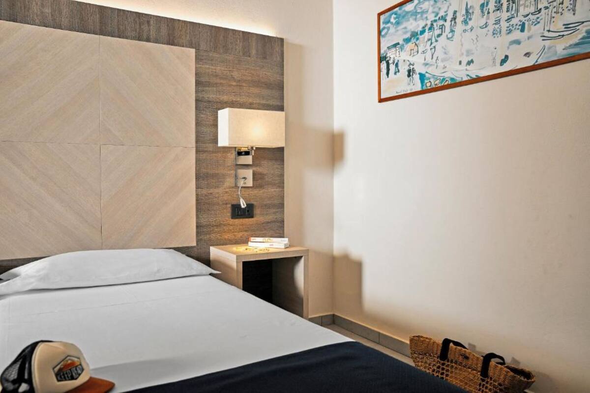 A hotel room with a bed, a nightstand with a lamp, a wall-mounted artwork, and a woven basket on the floor.