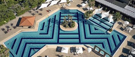 An aerial view of a resort with a large, geometric swimming pool, sun loungers, and shaded areas.