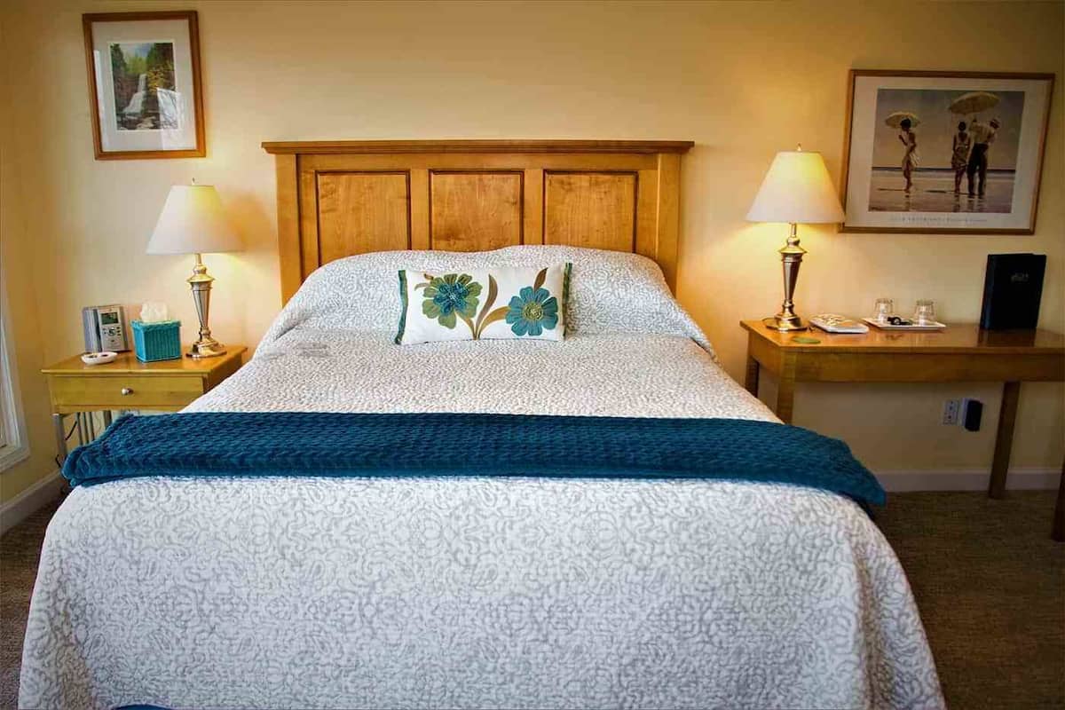 A neatly made bed with a floral pillow, two bedside lamps, a small table with a phone and a glass, and framed pictures on the walls.