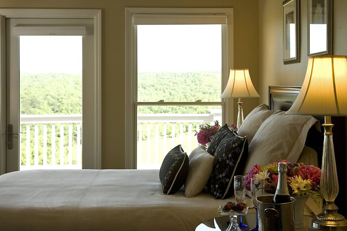 A hotel room with a bed, two bedside lamps, a nightstand with a vase of flowers, and a view of a balcony with a railing.