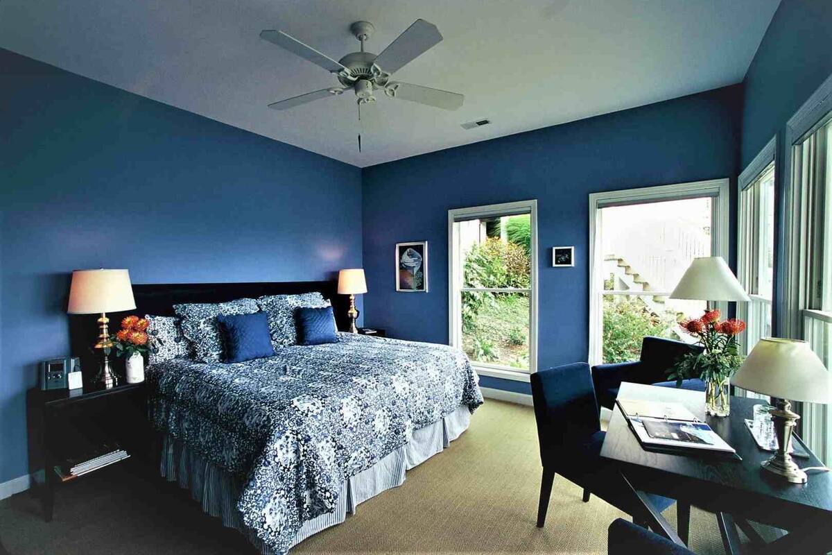 A bedroom with a bed, two bedside tables with lamps, a desk with a chair, a window, and a ceiling fan.