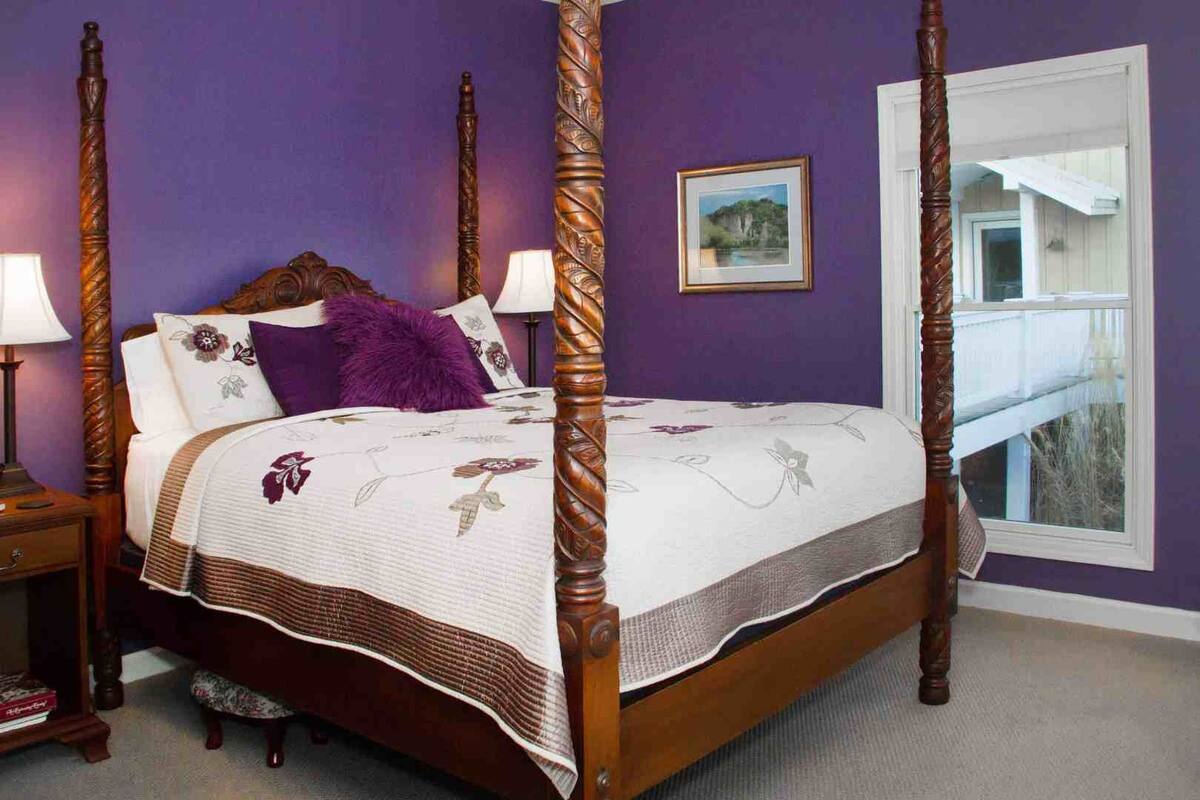 A four-poster bed with a floral bedspread, a purple throw pillow, and a framed picture on the wall.