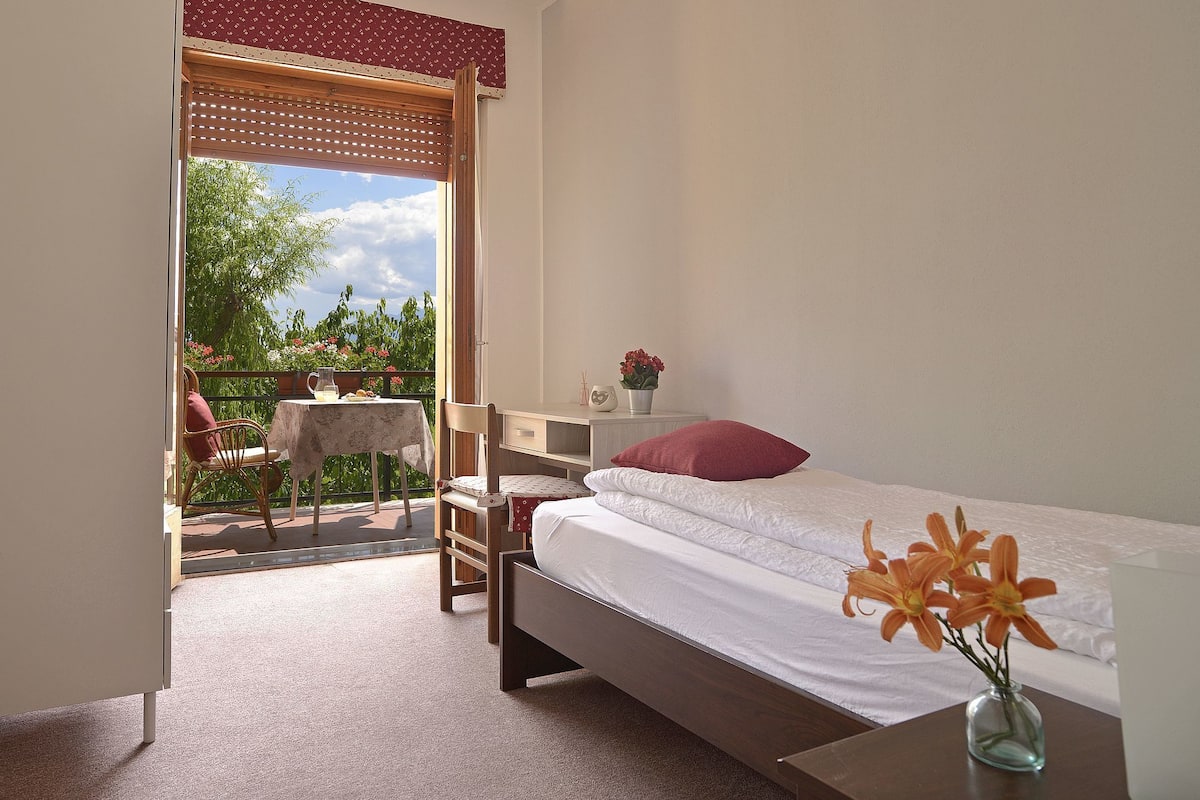 A bedroom with a bed, a small table, a chair, and a balcony with a table and chairs.