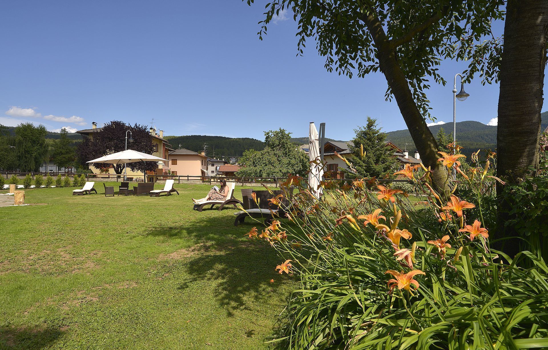 A grassy outdoor area with lounge chairs and a covered seating area, surrounded by trees and flowers.
