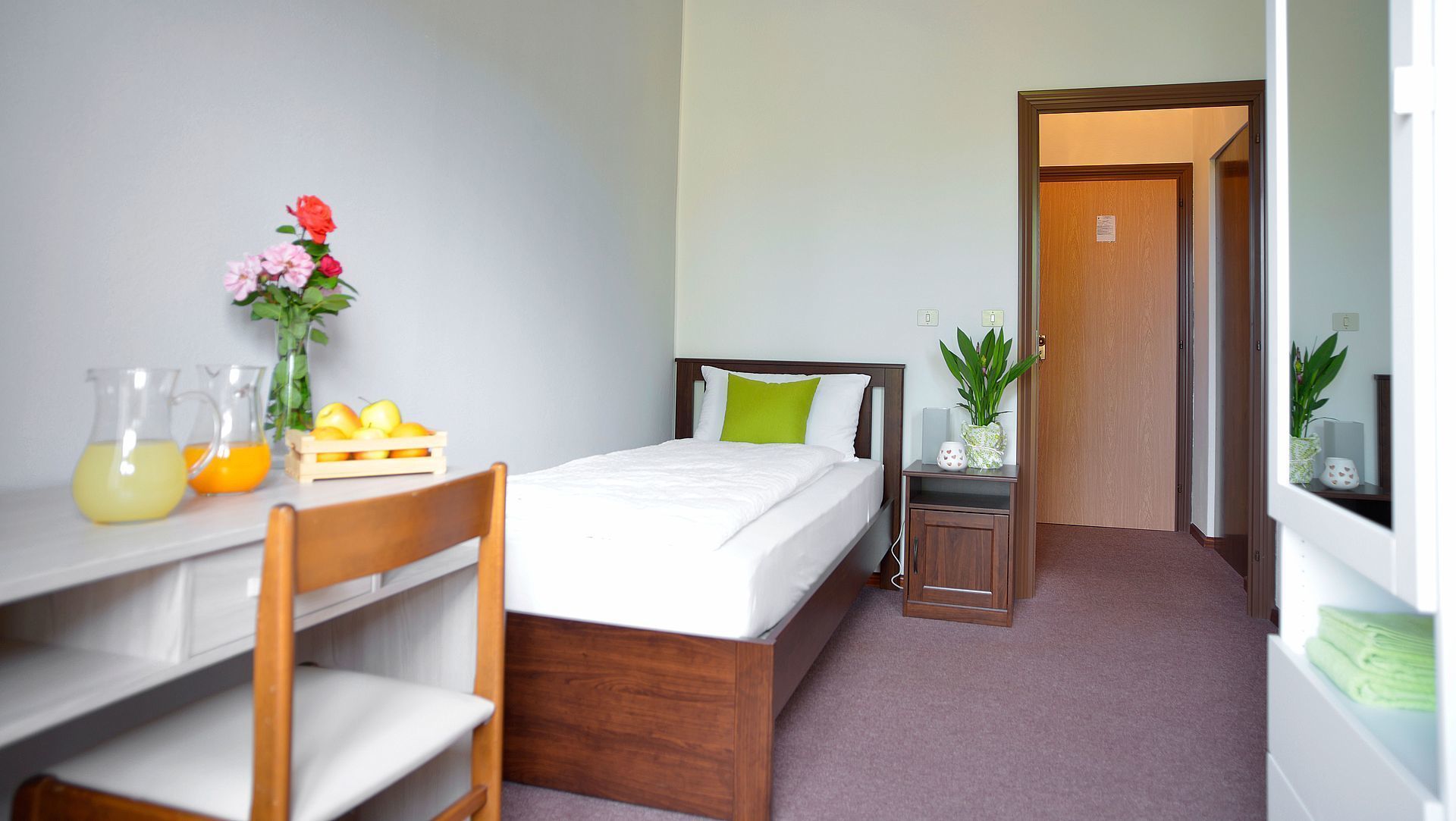 A small, neatly arranged bedroom with a single bed, a wooden nightstand, a desk with a chair, a small table with a vase of flowers, and a potted plant.