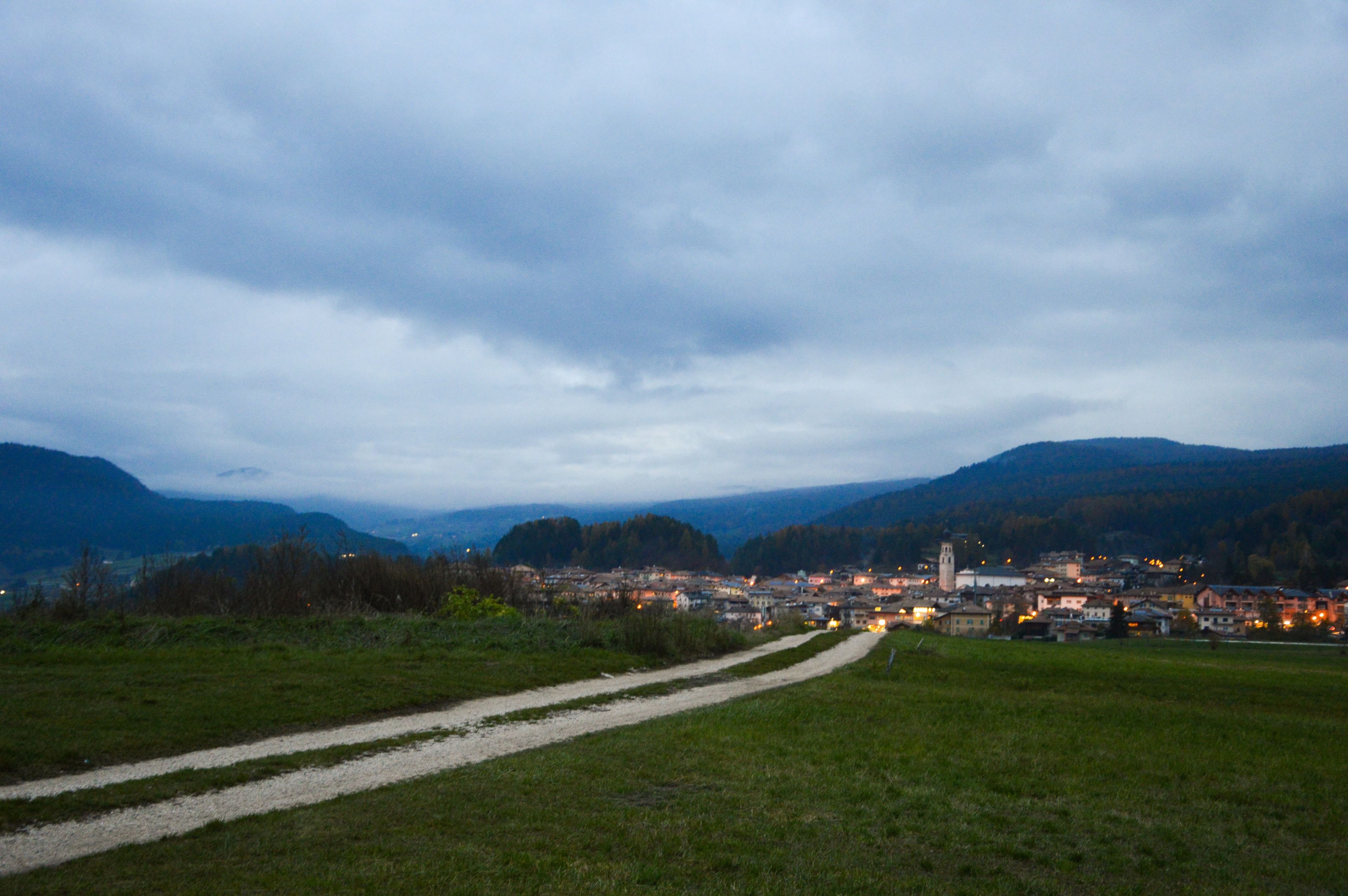 Un paese rurale con un sentiero sterrato che conduce a un gruppo di edifici, circondato da colline e un cielo nuvoloso.