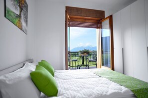 A bedroom with a bed, green pillows, a window with a view of mountains, and a balcony with a table and chairs. - Piccolo Orso Bruno (Cavareno)