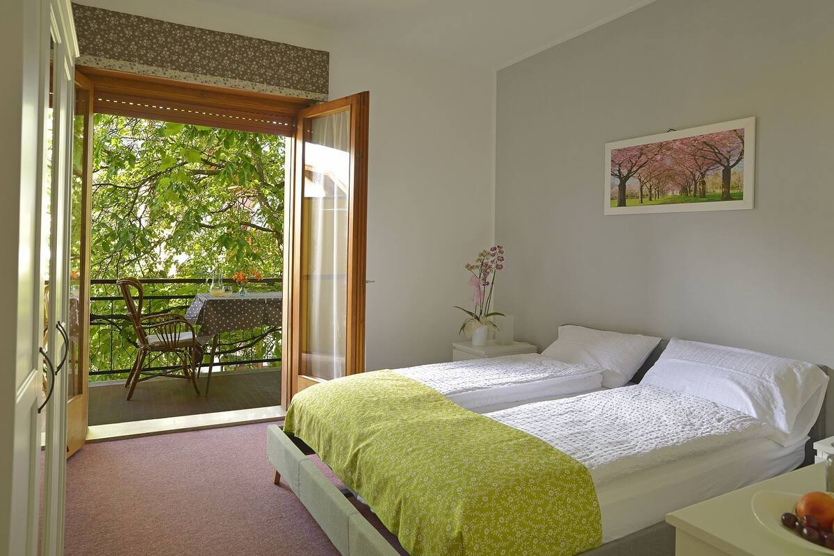 A bedroom with a bed, a small table with a vase and a picture, and a balcony with a chair and table.