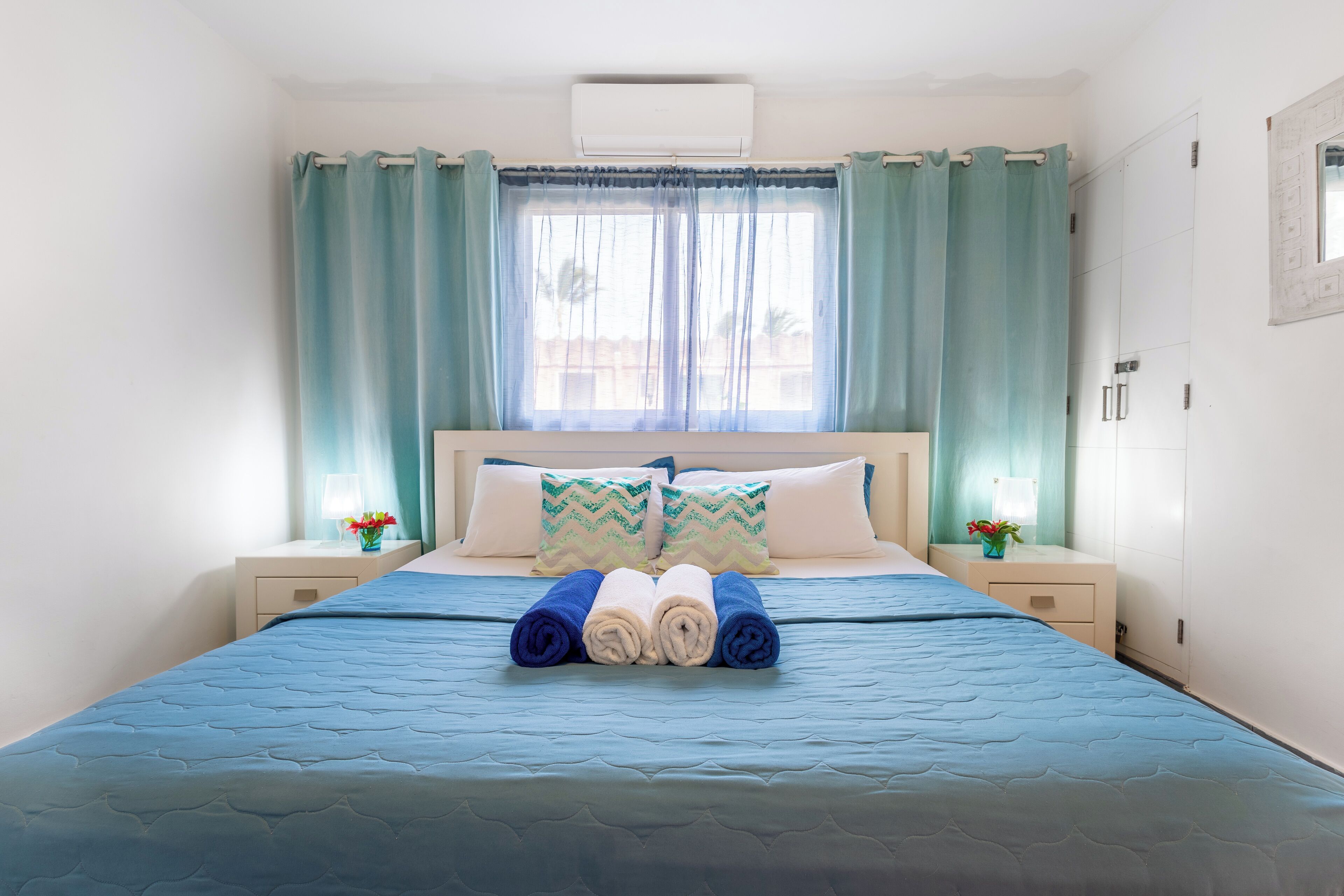 Una cama bien hecha con ropa de cama azul, dos almohadas y toallas enrolladas. Hay dos mesitas de noche con lámparas y jarrones de flores.