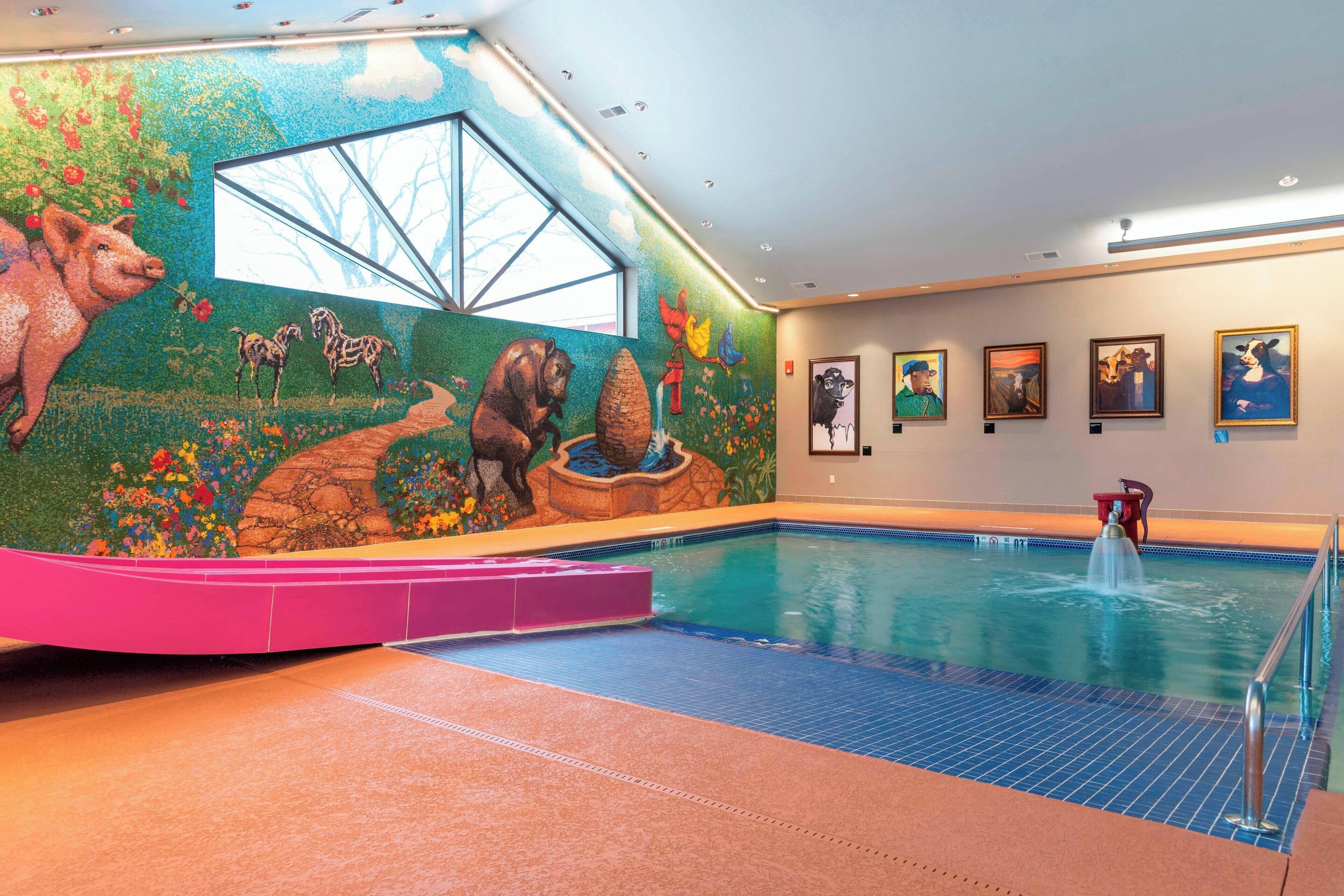 An indoor swimming pool with a mural of animals and flowers on the wall, a clear view of the sky through a window, and a water fountain.