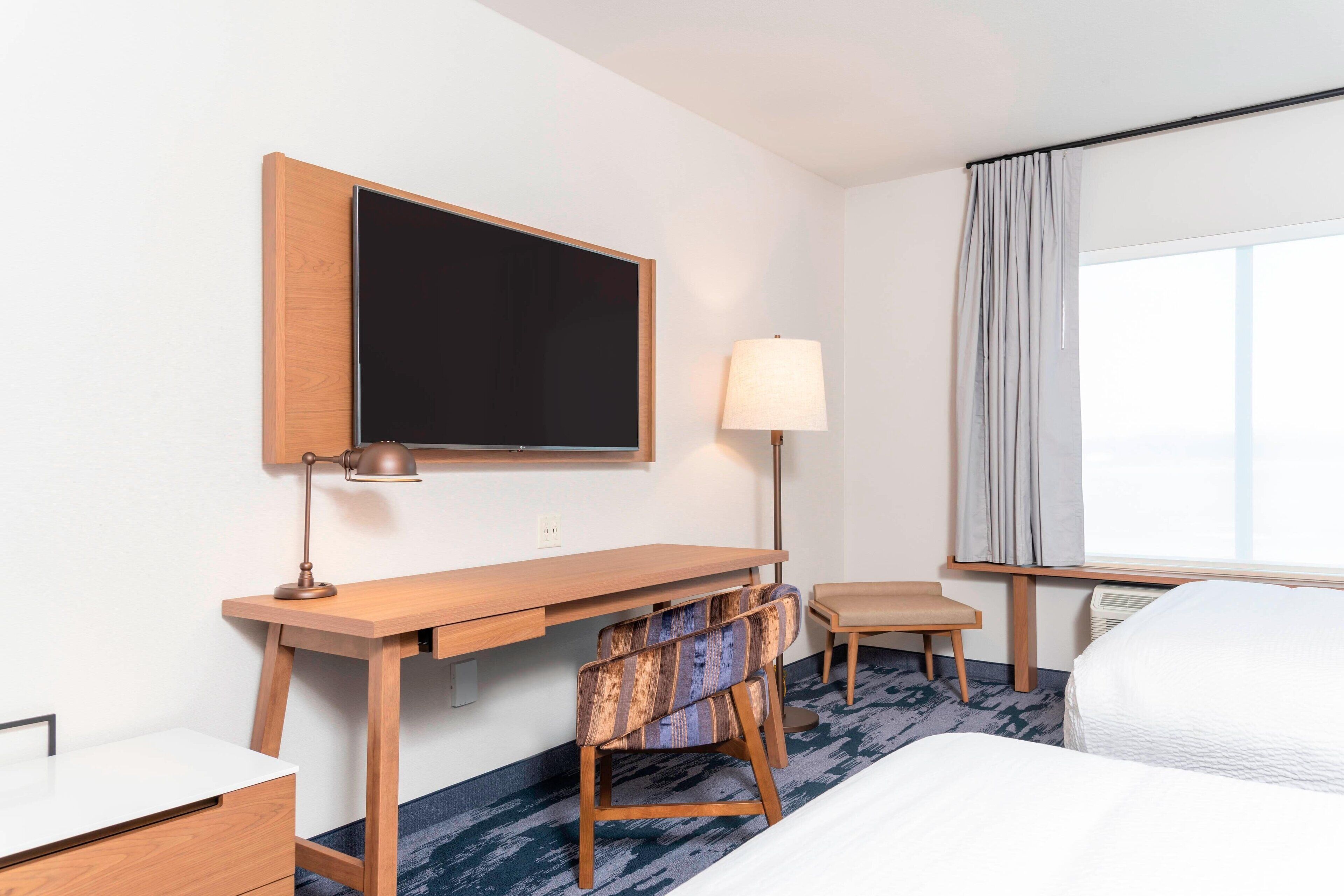 A hotel room with a wooden desk, a chair, a bed, a TV, and a window with curtains.